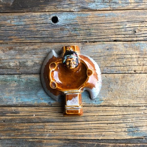 May include: A vintage ceramic ashtray shaped like an axe, with a Native American chief's face at the centre. The ashtray is mainly brown with grey and white details. It has a detailed design and is likely a collectable item.