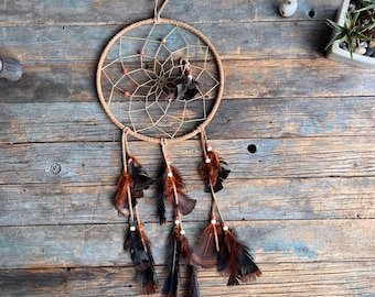 May include: A handcrafted dreamcatcher with a circular woven frame and a webbed center. Brown and black feathers dangle from the bottom, accented with small beads. The dreamcatcher is suspended against a rustic wooden background.
