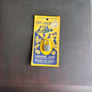 Puede incluir: Juego vintage de "Pet Lock" y "Luggage Lock" en una tarjeta amarilla y azul. La tarjeta presenta ilustraciones de perros, equipaje y un candado. El candado es plateado con las letras "SLC". La tarjeta también tiene el texto "a prueba de óxido".