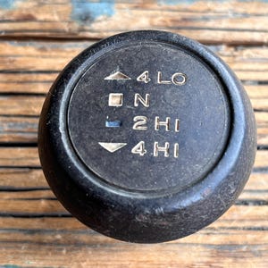 May include: A black, circular gear shift knob with white text and directional indicators. The text includes "4 LO", "N", "2 HI", and "4 HI". The knob is set against a weathered, wooden surface.