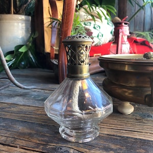 Könnte beinhalten: Eine klare Glas-Öllampe mit einem Messingdeckel und einem Docht. Die Lampe steht auf einer Holzoberfläche.