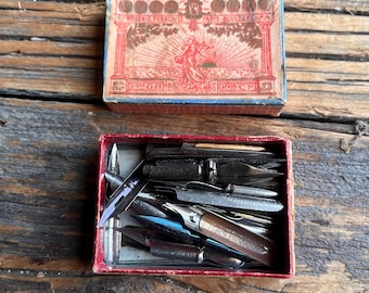 Könnte beinhalten: Eine vintage rote Schachtel gefüllt mit einer Vielzahl von antiken Füllfederhalterspitzen. Die Schachtel ist mit "Compagnie Française Paris Boulogne-sur-Seine" beschriftet.