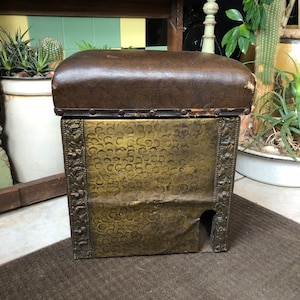May include: A vintage, gold-colored metal footstool with a brown leather top. The stool has a decorative embossed pattern and a cutout on the front.