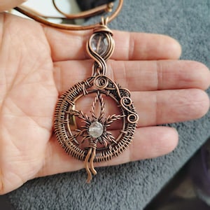 May include: A copper wire-wrapped pendant featuring a circular frame with a central leaf-shaped design and two clear faceted gemstones. The pendant is suspended from a brown leather cord and showcases intricate metalwork with coiled accents.