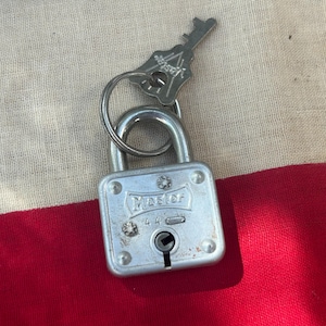 May include: A silver-colored Master padlock with a key attached by a metal ring. The key has a decorative design. The padlock is on a red and white striped background.