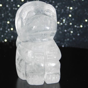 May include: A clear quartz crystal carving of a stylized owl. The owl is depicted in a seated position with its wings folded. The carving is detailed, with visible features such as eyes, beak, and feathers. The crystal is translucent.
