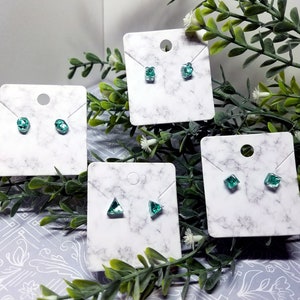 May include: Four pairs of earrings displayed on marble cards. The earrings are all different shapes and sizes, and they are all a light green color.