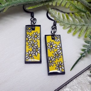May include: A pair of rectangular earrings with a yellow background and a daisy pattern. The daisies are white with black outlines. The earrings have black metal frames and ear wires.
