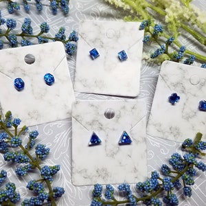May include: Four cards with blue gemstone earrings on white marble backgrounds. The earrings are in different shapes, including square, round, flower, and triangle.