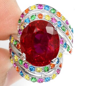 May include: A silver ring featuring a large red oval gemstone encircled by a rainbow of smaller gemstones.