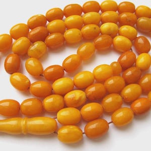 Puede incluir: Un collar de cuentas de color ámbar. Las cuentas son de forma ovalada y están ensartadas, con una cuenta más grande y alargada en el extremo. El color de las cuentas varía del amarillo claro a un marrón anaranjado más oscuro.