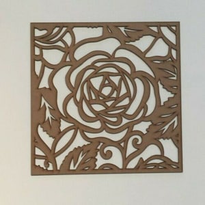 May include: A square, laser-cut wooden panel featuring a rose design. The panel is a warm brown color, with the rose and surrounding leaves and vines cut out, creating a negative space. The design is centered on the panel.