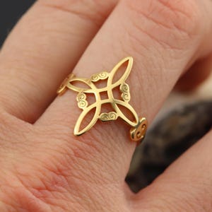 May include: A gold-colored ring with an openwork design featuring a central square and swirling details. The ring is worn on a finger, showcasing its intricate pattern and metallic sheen. The ring's design is symmetrical and ornate.