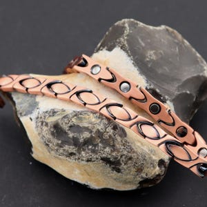 May include: Two copper magnetic bracelets with a wave-like design. The bracelets are stacked on top of each other and have silver-colored magnetic closures.