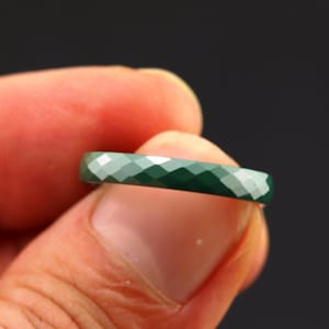 Puede incluir: Un anillo facetado de color verde oscuro se sostiene entre los dedos sobre un fondo oscuro. La superficie del anillo está cortada con múltiples facetas, creando un efecto brillante. El anillo parece estar hecho de una piedra preciosa o material similar.
