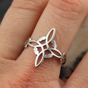 Könnte beinhalten: Ein silberner Ring mit einem filigranen Design, das ein zentrales Quadrat und vier längliche, gebogene Elemente aufweist. Der Ring hat dekorative Wirbel an den Seiten. Der Ring wird an einem Finger getragen.