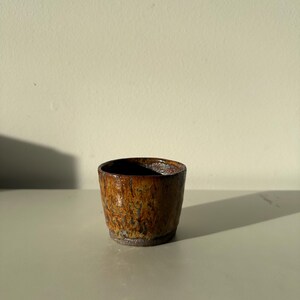 May include: A small, handcrafted ceramic cup with a textured, mottled brown glaze. The cup has a dark interior and a slightly flared rim. The exterior shows variations in colour, from light brown to dark brown, with hints of orange.