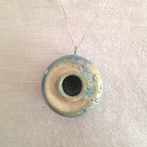 May include: A ceramic pendant necklace with a light blue and white glaze. The pendant is shaped like a donut with a hole in the center. The pendant has a snowflake design.