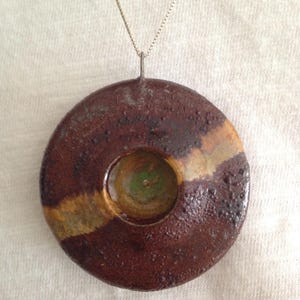 May include: A round ceramic pendant necklace with a brown, tan, and green glaze. The pendant has a small hole in the center.