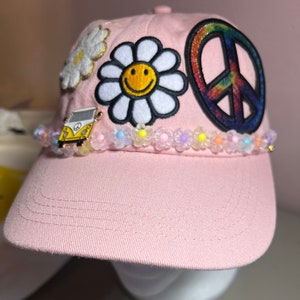 May include: Pink baseball cap with a peace sign, daisy, and a yellow Volkswagen van patch. The cap is decorated with a row of colourful flower beads.