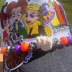 Puede incluir: Gorra de béisbol blanca con visera negra y parches bordados de colores con las hermanas Sanderson de la película Hocus Pocus. La gorra también tiene un dije de calavera y una cadena de cuentas con cuentas naranjas, moradas y negras.