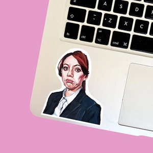 May include: A sticker of a woman with red hair wearing a dark blue blazer and a white shirt. She has a serious expression on her face.