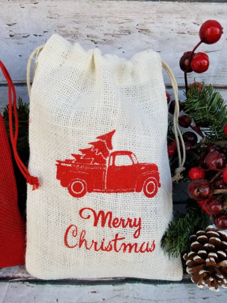 Pu&ograve; includere: Un sacchetto di iuta color crema con coulisse e un camion rosso vintage che trasporta un albero di Natale. Le parole "Merry Christmas" sono stampate in rosso sotto. Il sacchetto ha un cordoncino color naturale.