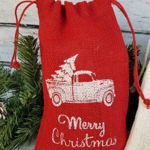 Pu&ograve; includere: Un sacchetto rosso in iuta con coulisse e una grafica bianca di un camion vintage che trasporta un albero di Natale. Le parole "Merry Christmas" sono stampate in bianco. Sacchetto regalo natalizio.
