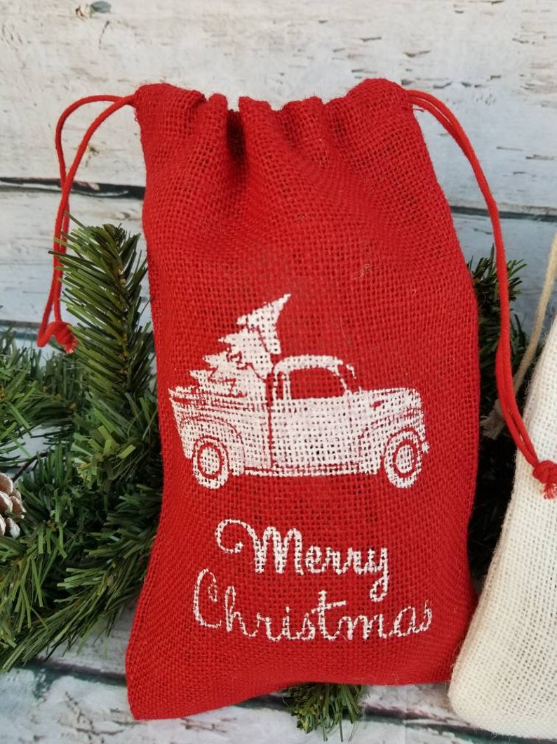 Pu&ograve; includere: Un sacchetto rosso in iuta con coulisse e una grafica bianca di un camion vintage che trasporta un albero di Natale. Le parole "Merry Christmas" sono stampate in bianco. Sacchetto regalo natalizio.