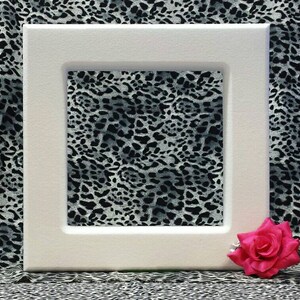 May include: A white picture frame with a black and white animal print design inside. A pink flower is sitting on the bottom right corner of the frame.