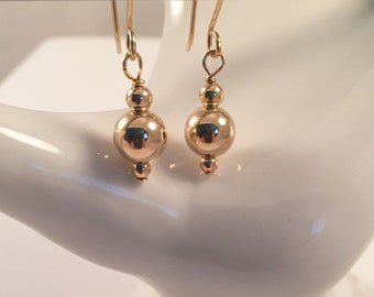 May include: A pair of gold-colored earrings. Each earring features a small, round bead at the top, connected to a larger, polished sphere. The earrings hang from gold-colored ear wires. The earrings are displayed against a white background.