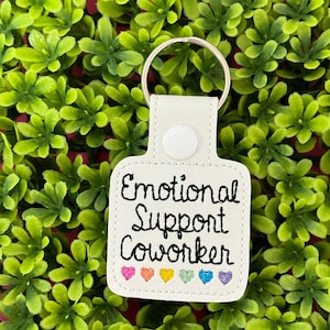 May include: White leatherette keychain with the text "Emotional Support Coworker" in black script. The keychain features embroidered hearts in rainbow colors. A silver key ring is attached.
