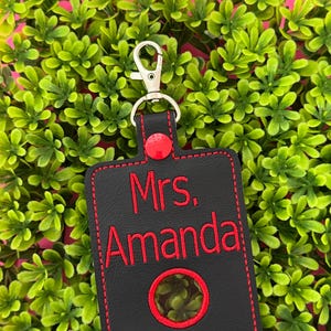 May include: Black rectangular keychain with red stitching and a silver clasp. The keychain features the embroidered text "Mrs. Amanda" in red. A red circle is embroidered at the bottom. The background is a bed of green leaves.