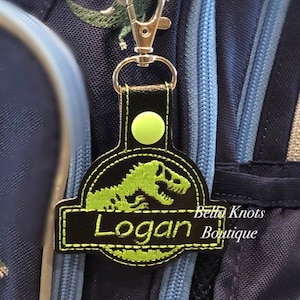 May include: A personalized black and green dinosaur-themed keychain with the name "Logan" embroidered on it. The keychain is attached to a navy blue backpack with dinosaur embroidery.