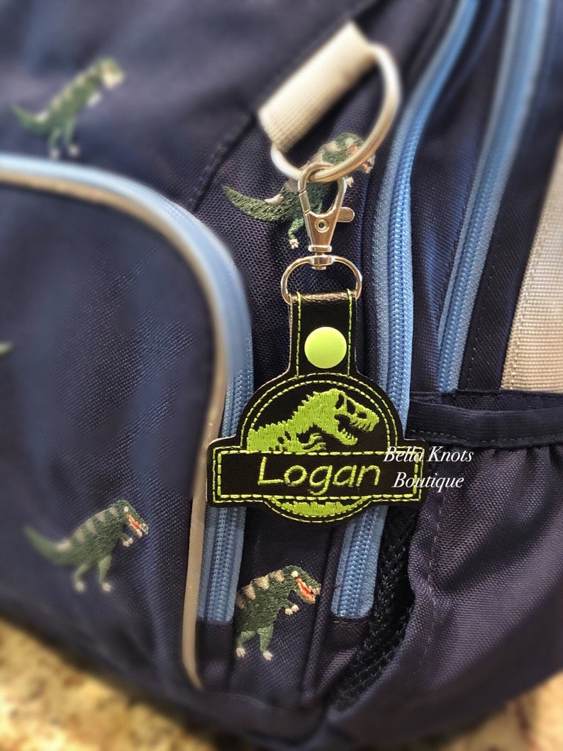 May include: A personalized black and green dinosaur-themed keychain with the name "Logan" embroidered on it. The keychain is attached to a navy blue backpack with dinosaur embroidery.
