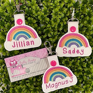 May include: Three rainbow-themed keychains with personalized names: Jillian, Sadey, and Magnus J. Each keychain features a white background with a colorful rainbow and a pink button.