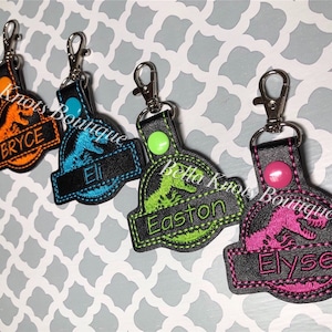 May include: Embroidered keychains featuring a dinosaur design with names. Each keychain has a different color scheme: red, orange, blue, green, and pink. The names Ayden, Bryce, Eli, Easton, and Elyse are embroidered on the keychains.