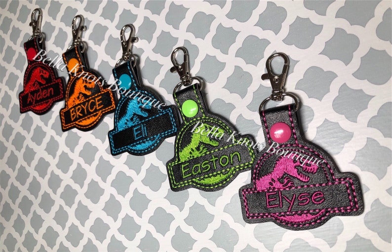 May include: Embroidered keychains featuring a dinosaur design with names. Each keychain has a different color scheme: red, orange, blue, green, and pink. The names Ayden, Bryce, Eli, Easton, and Elyse are embroidered on the keychains.
