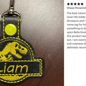 May include: Black and yellow embroidered keychain with a dinosaur design and the name "Liam". The keychain has a metal clasp and a yellow button. The text "Choose Thread Color: Yellow" is also visible.