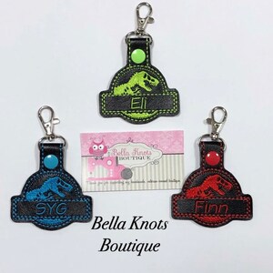 May include: Three keychains with a dinosaur design. One is green with "Eli", one is blue with "SYG", and one is red with "Finn". Each keychain has a black background and a silver clasp. The Bella Knots Boutique logo is also visible.