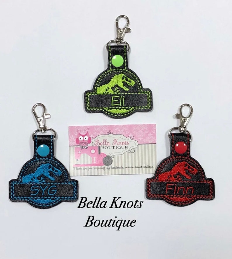 May include: Three keychains with a dinosaur design. One is green with "Eli", one is blue with "SYG", and one is red with "Finn". Each keychain has a black background and a silver clasp. The Bella Knots Boutique logo is also visible.