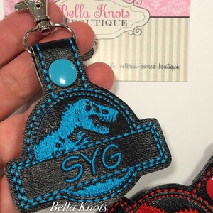 May include: Black keychain with a blue dinosaur design and the letters "SYG". The keychain has a silver clasp and a blue button. The text "Bella Knots Boutique" is visible on the keychain.