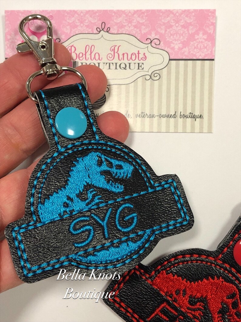 May include: Black keychain with a blue dinosaur design and the letters "SYG". The keychain has a silver clasp and a blue button. The text "Bella Knots Boutique" is visible on the keychain.