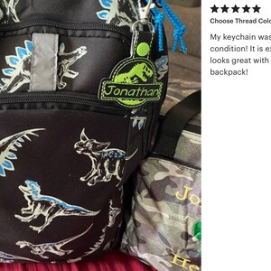 May include: Black backpack with dinosaur print, featuring a reflective strip and a blue zipper. Includes a personalized keychain with the name "Jonathan" and a small plush dinosaur. A matching lunch bag is also visible.