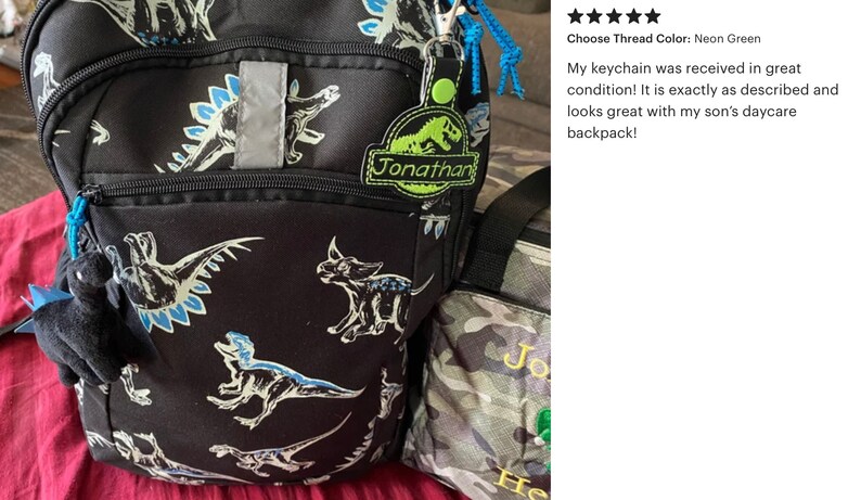 May include: Black backpack with dinosaur print, featuring a reflective strip and a blue zipper. Includes a personalized keychain with the name "Jonathan" and a small plush dinosaur. A matching lunch bag is also visible.