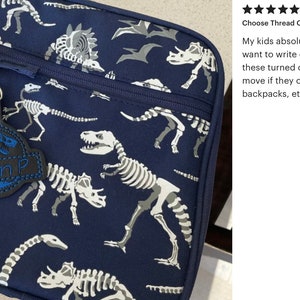 May include: Navy blue lunch bag with a dinosaur skeleton pattern. A black and blue tag with the name "Liam P" is attached to the zipper. The bag features a zipper closure and a side mesh pocket.