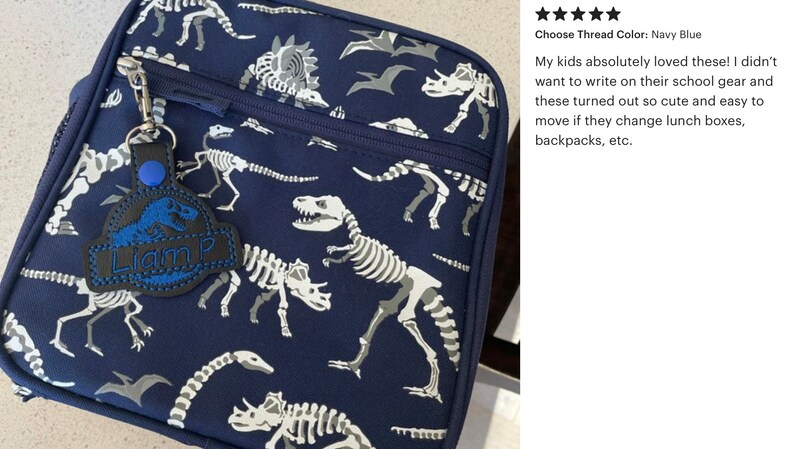 May include: Navy blue lunch bag with a dinosaur skeleton pattern. A black and blue tag with the name "Liam P" is attached to the zipper. The bag features a zipper closure and a side mesh pocket.