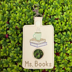 May include: Cream-colored keychain with a silver clasp. The keychain features an embroidered design of a steaming green mug atop a stack of books. The text "Ms. Books" is embroidered below the books.