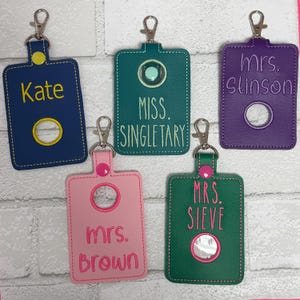 May include: Five rectangular keychains with metal lobster clasps, featuring embroidered names and circular cutouts. The colors include navy blue with yellow text, teal with light green text, purple with purple text, pink with pink text, and green with pink text.
