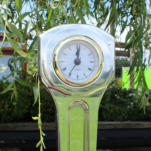 May include: A silver clock shaped like a car engine connecting rod. The clock face is white with black numbers and hands. The clock is sitting on a wooden base.